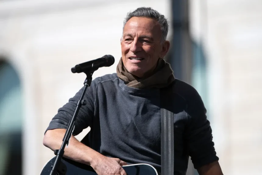 A man with short gray hair smiles warmly while holding a guitar and standing at a microphone outdoors, wearing a dark sweater and olive scarf