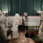 A tattooed figure watches a wedding ceremony beside an open casket surrounded by flowers.