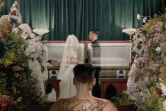 A tattooed figure watches a wedding ceremony beside an open casket surrounded by flowers.