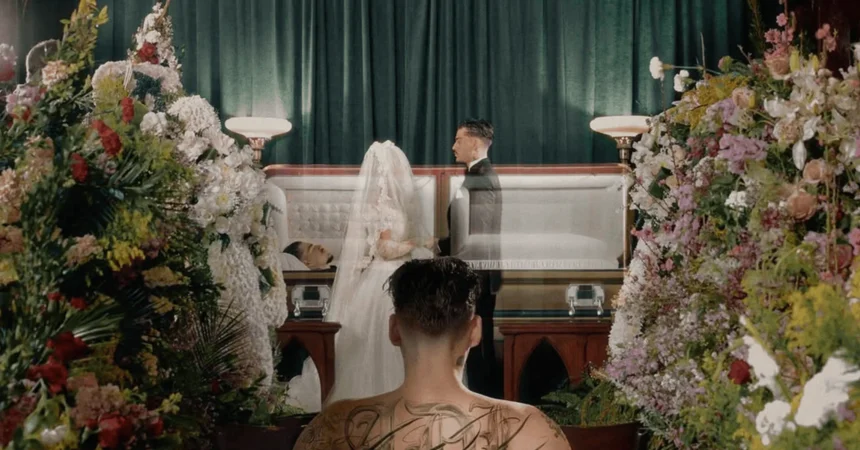 A tattooed figure watches a wedding ceremony beside an open casket surrounded by flowers.