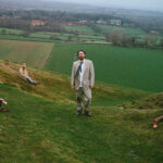 Gently Tender band members posed across a green English hillside, fields and grey sky behind them.