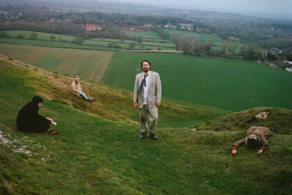 Gently Tender band members posed across a green English hillside, fields and grey sky behind them.