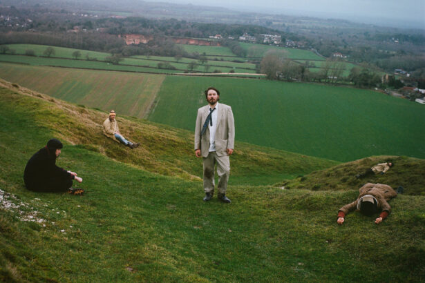 Gently Tender band members posed across a green English hillside, fields and grey sky behind them.