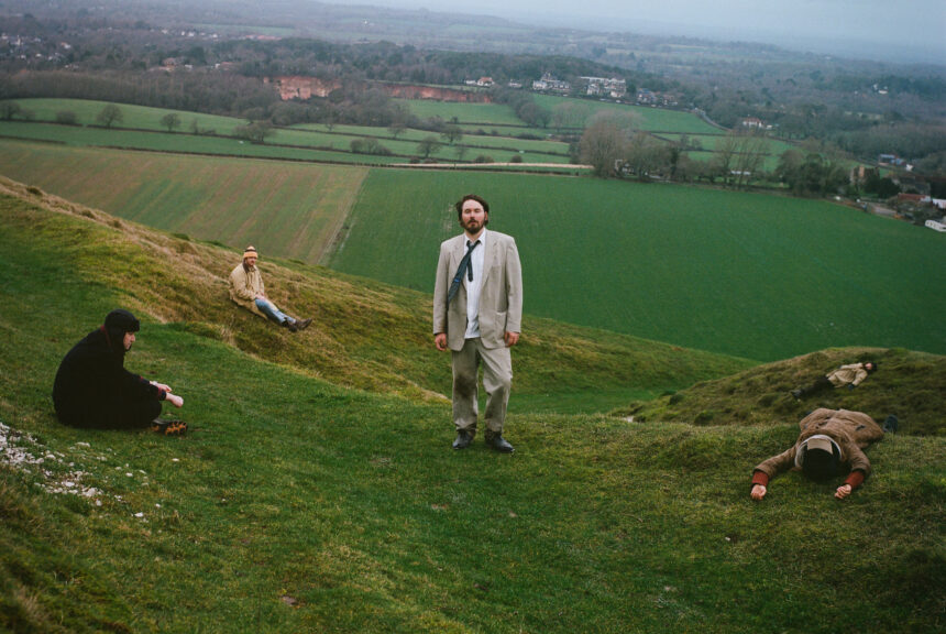 Gently Tender band members posed across a green English hillside, fields and grey sky behind them.