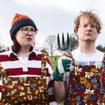Joel X Eleanor standing in a park wearing colorful overalls, holding a garden fork