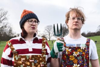 Joel X Eleanor standing in a park wearing colorful overalls, holding a garden fork
