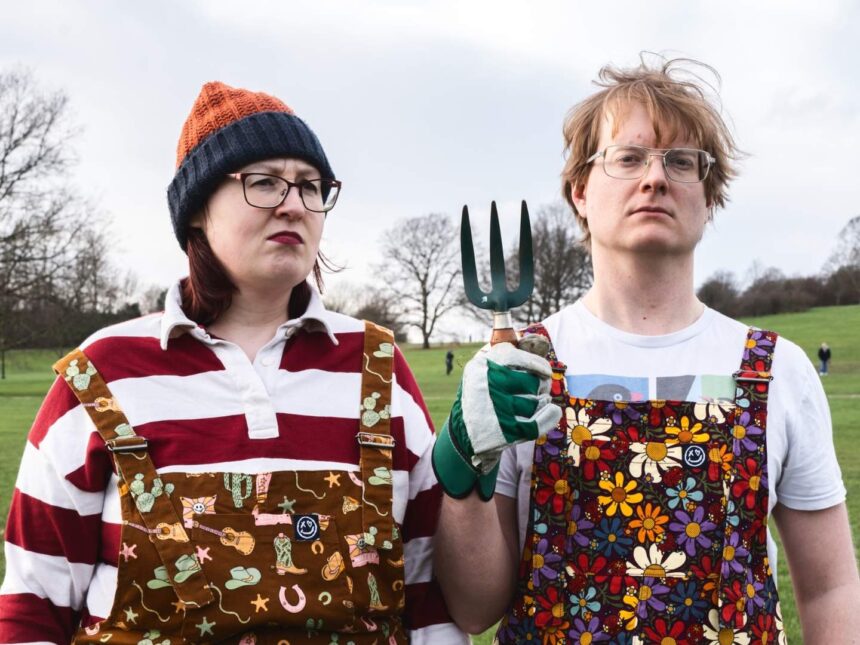 Joel X Eleanor standing in a park wearing colorful overalls, holding a garden fork