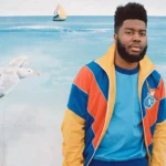Singer Khalid wearing a colorful Gucci track jacket, posed in front of an ocean mural.