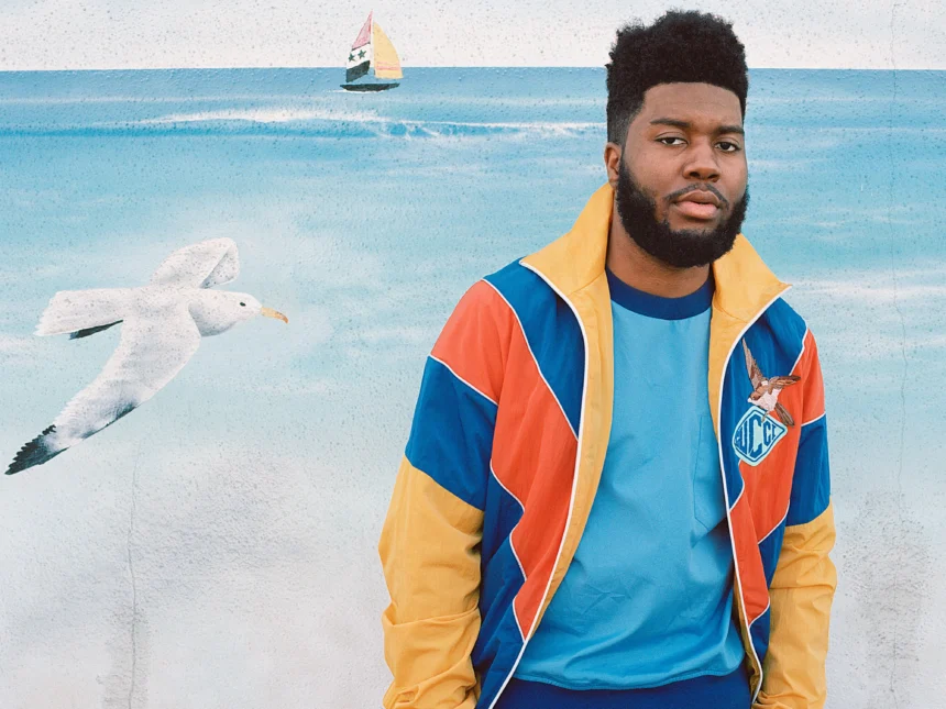 Singer Khalid wearing a colorful Gucci track jacket, posed in front of an ocean mural.