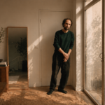 A candid indoor portrait of Thomas Bangalter standing by a sunlit window, dressed casually in a dark sweater and loose trousers, surrounded by warm wooden textures and soft natural light.