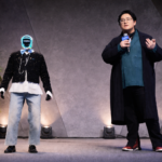 A presenter holding a microphone speaks on stage alongside a humanoid robot at a tech event.