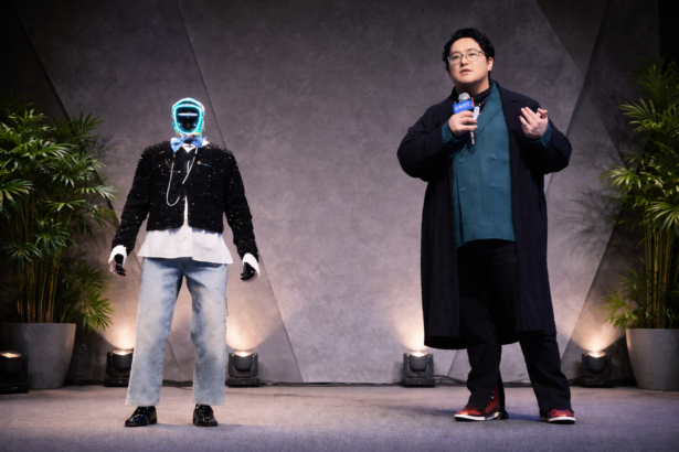 A presenter holding a microphone speaks on stage alongside a humanoid robot at a tech event.