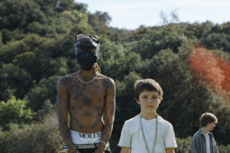 A shirtless tattooed person in a mask stands beside a young boy on a grassy hillside, with a third figure visible in the background.