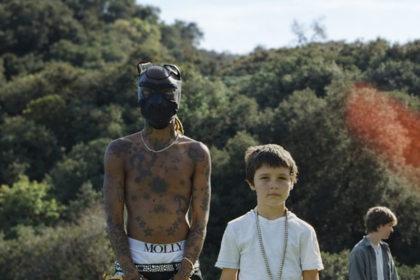 A shirtless tattooed person in a mask stands beside a young boy on a grassy hillside, with a third figure visible in the background.