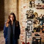 Belle Blue stands in a brick-walled studio space surrounded by vintage rock posters, a guitar, vinyl records, and a Marshall amp, wearing a navy coat and grey scarf.