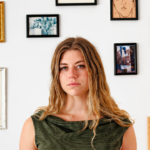Ella Clayton standing before a gallery wall of framed artwork, promoting her second album 'Could It Be You?'