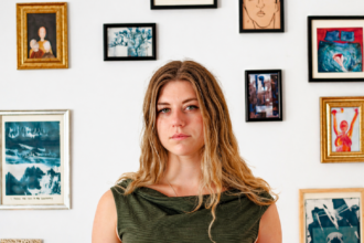 Ella Clayton standing before a gallery wall of framed artwork, promoting her second album 'Could It Be You?'