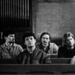 Basht sit in a dim, church-like space as they announce their debut album Poor Advice, arriving in October, alongside the lead single “Perfume.”