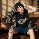 Chloe Star poses on a leather couch wearing a Miami Alchemist tee, leopard jacket, denim shorts, and black cap, with tattoos visible throughout.