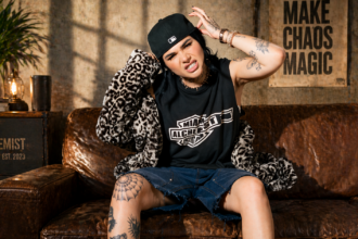 Chloe Star poses on a leather couch wearing a Miami Alchemist tee, leopard jacket, denim shorts, and black cap, with tattoos visible throughout.
