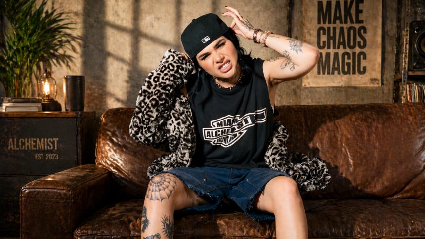 Chloe Star poses on a leather couch wearing a Miami Alchemist tee, leopard jacket, denim shorts, and black cap, with tattoos visible throughout.
