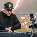 DJ Shadow performing live on stage at an outdoor festival, focused over Pioneer DJ decks and Serato setup, wearing all black and a Golden State Warriors cap.