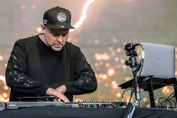DJ Shadow performing live on stage at an outdoor festival, focused over Pioneer DJ decks and Serato setup, wearing all black and a Golden State Warriors cap.