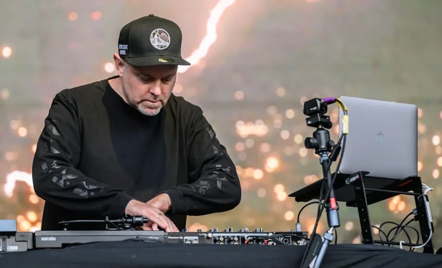 DJ Shadow performing live on stage at an outdoor festival, focused over Pioneer DJ decks and Serato setup, wearing all black and a Golden State Warriors cap.