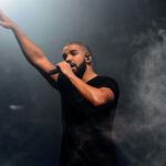Drakeon stage holding a microphone and raising one arm, surrounded by dramatic smoke and spotlight lighting, wearing a black t-shirt.