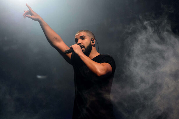 Drakeon stage holding a microphone and raising one arm, surrounded by dramatic smoke and spotlight lighting, wearing a black t-shirt.