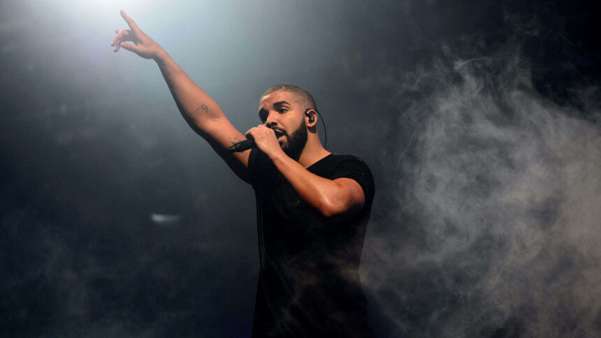 Drakeon stage holding a microphone and raising one arm, surrounded by dramatic smoke and spotlight lighting, wearing a black t-shirt.