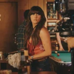 Ella Langley stands at a bar in a music video still, wearing a red fringe top and embroidered denim shorts, with a pool table and honky-tonk interior visible behind her.