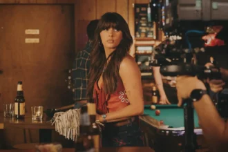 Ella Langley stands at a bar in a music video still, wearing a red fringe top and embroidered denim shorts, with a pool table and honky-tonk interior visible behind her.