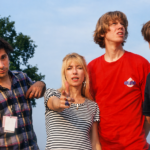 Four members of Sonic Youth posing outdoors, with one pointing at the camera and others looking in different directions under a bright sky.