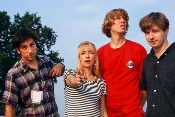 Four members of Sonic Youth posing outdoors, with one pointing at the camera and others looking in different directions under a bright sky.