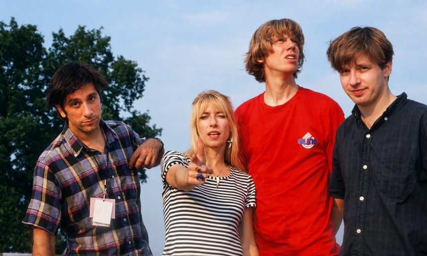 Four members of Sonic Youth posing outdoors, with one pointing at the camera and others looking in different directions under a bright sky.