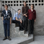 Four young men posed on concrete outdoor stairs against a weathered grey wall, wearing casual button-up shirts, with serious expressions - LateTown