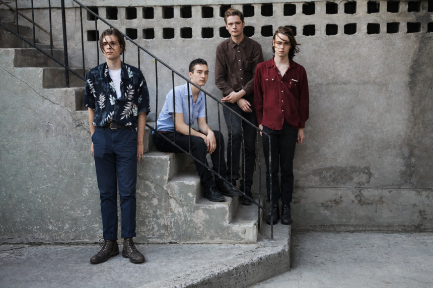 Four young men posed on concrete outdoor stairs against a weathered grey wall, wearing casual button-up shirts, with serious expressions - LateTown
