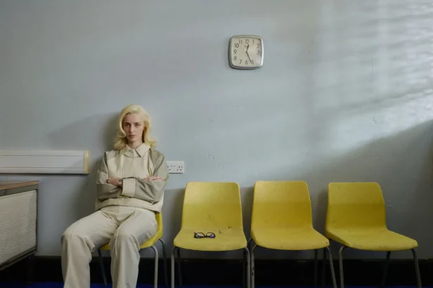 `Gia Ford seated alone in a waiting room among yellow chairs, arms crossed, capturing the glum and unmoored mood of her new single 'I Guess My Time Has Come'.`