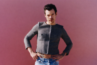 Harry Styles Man posing confidently against a pink background, wearing a cropped jacket and jeans