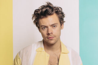 Harry Styles poses in a soft-lit studio portrait wearing a sheer yellow-and-white shirt against a pastel backdrop.