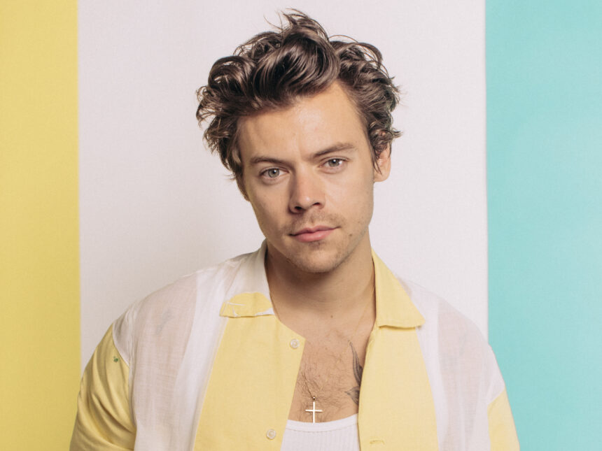 Harry Styles poses in a soft-lit studio portrait wearing a sheer yellow-and-white shirt against a pastel backdrop.
