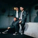 Holding Absence trio poses in an industrial setting wearing matching dark workwear, promoting their 2026 single 'Whisper Of A Dream' on Sumerian Records