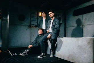 Holding Absence trio poses in an industrial setting wearing matching dark workwear, promoting their 2026 single 'Whisper Of A Dream' on Sumerian Records