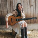 Kacey Musgraves sits on a wooden stump in a barn, holding an acoustic guitar, wearing a cream knit sweater and dark brown knee-high leather boots, with hay on the floor behind her - LateTown