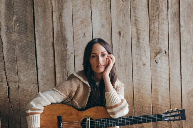 Kacey Musgraves sits on a wooden stump in a barn, holding an acoustic guitar, wearing a cream knit sweater and dark brown knee-high leather boots, with hay on the floor behind her - LateTown