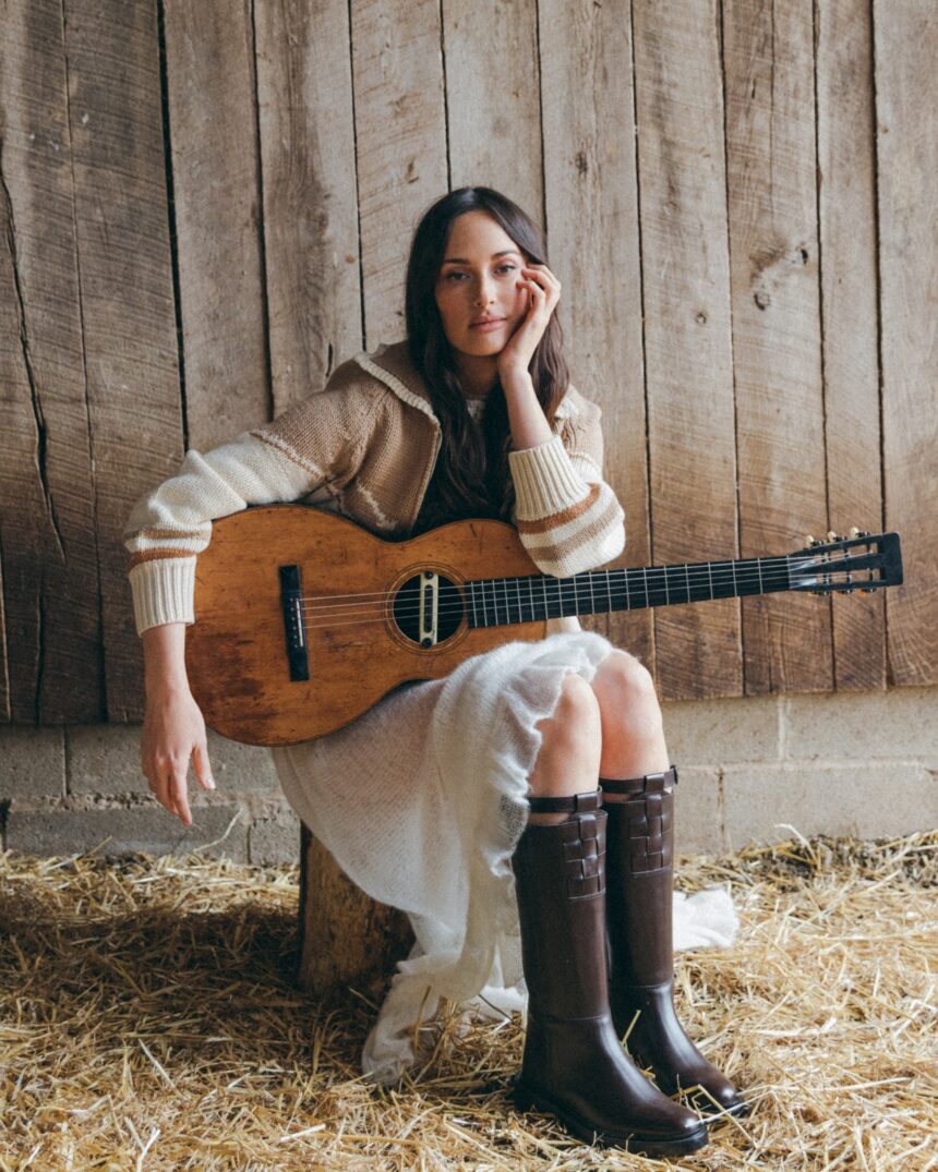 Kacey Musgraves sits on a wooden stump in a barn, holding an acoustic guitar, wearing a cream knit sweater and dark brown knee-high leather boots, with hay on the floor behind her - LateTown
