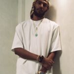 Keem wearing an all-white outfit, gold chain with green pendant, bandana headwrap, and a gold watch, leaning against an arched wall.