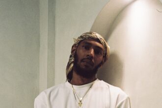Keem wearing an all-white outfit, gold chain with green pendant, bandana headwrap, and a gold watch, leaning against an arched wall.