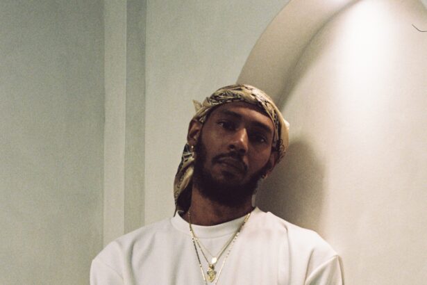 Keem wearing an all-white outfit, gold chain with green pendant, bandana headwrap, and a gold watch, leaning against an arched wall.